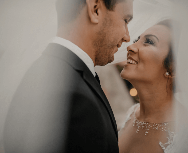 a close-up of a bride and groom smiling at each other, about to kiss. The bride's veil is flowing around them.